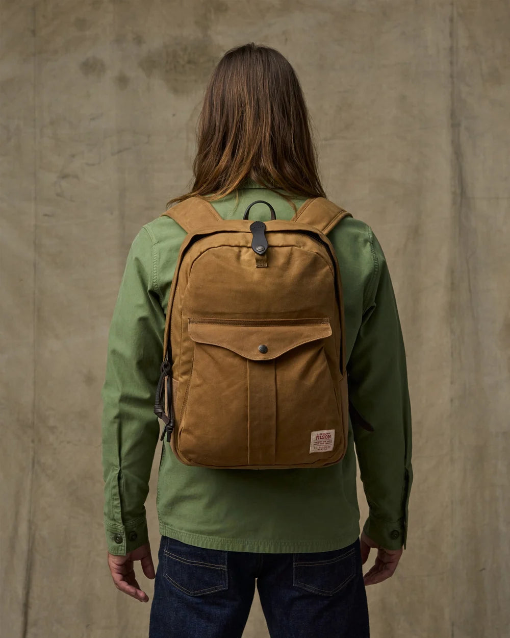 Person wearing a brown backpack and green jacket against a beige wall