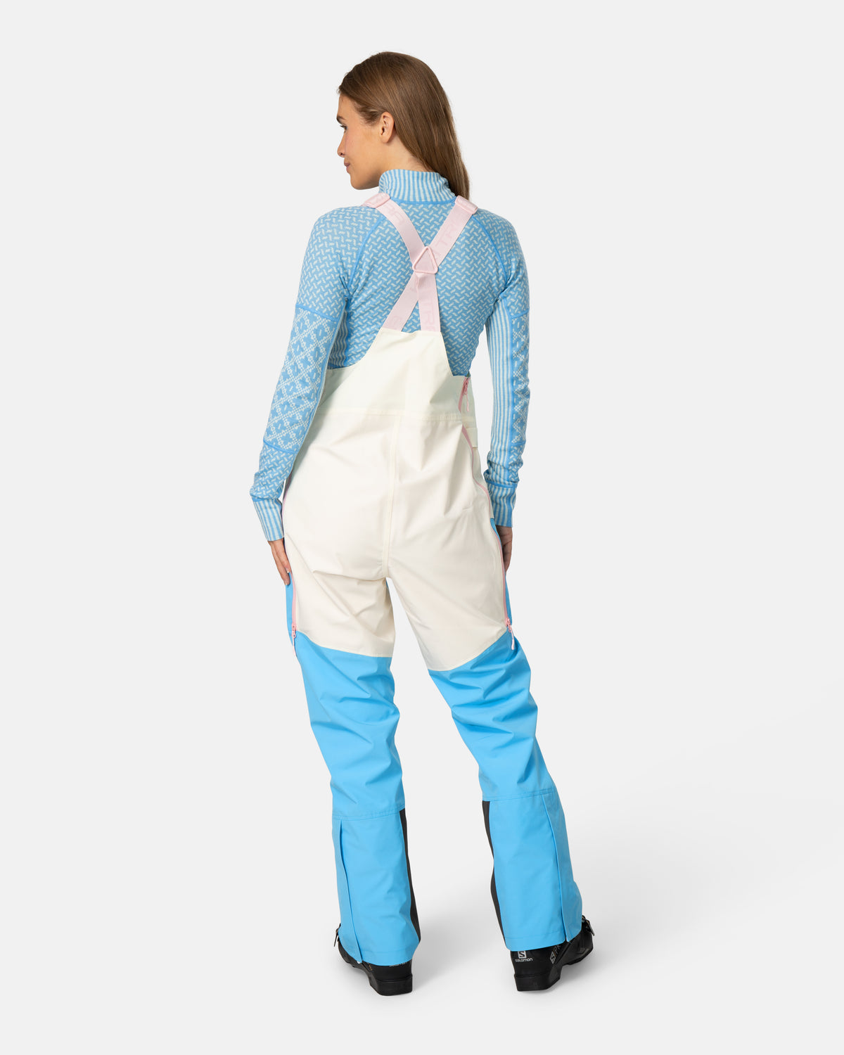 A model shown from the back wearing a color-blocked ski bib over a light blue patterned long-sleeve shirt. The bib features an off-white upper section and bright blue lower legs, with light pink adjustable shoulder straps.