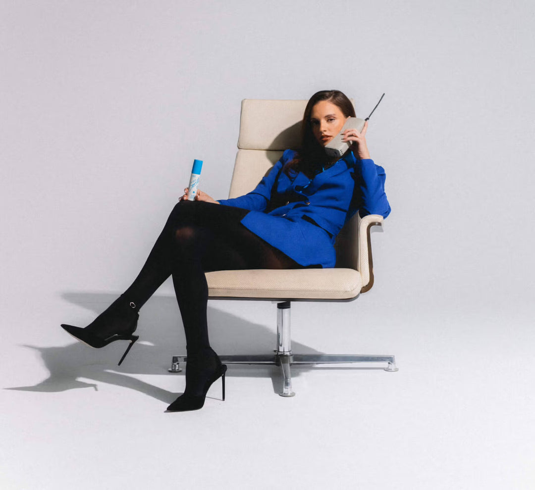 Woman in a blue coat sitting on a chair holding a phone and a can, against a white background