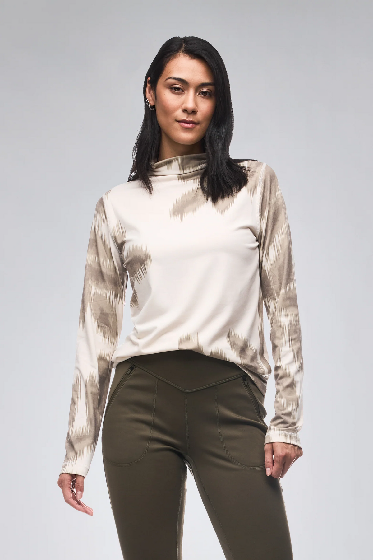 A woman modeling a long-sleeve athletic top with a beige and taupe tie-dye pattern and olive green leggings, standing against a light gray studio background.