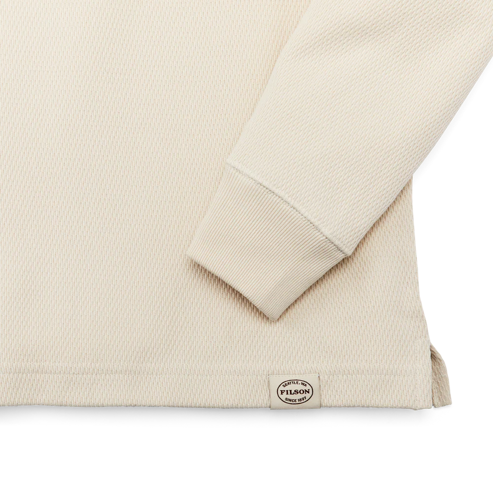 Close-up of a light beige ribbed knit long-sleeve top showing the sleeve cuff and a small circular Piltdown brand tag near the hem, on a white background.