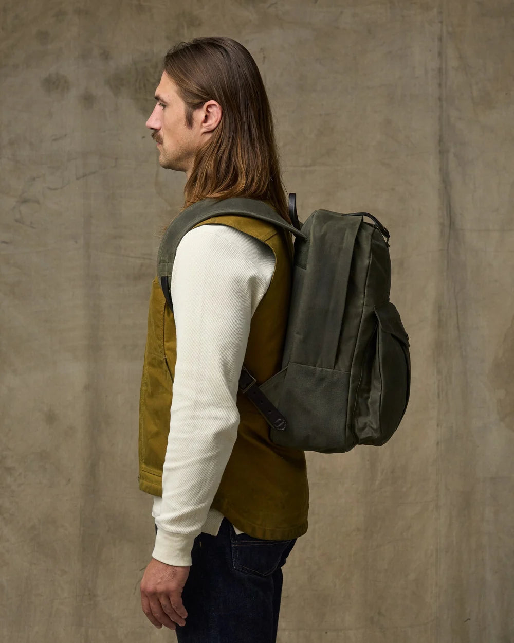 Man wearing a green backpack against a beige wall