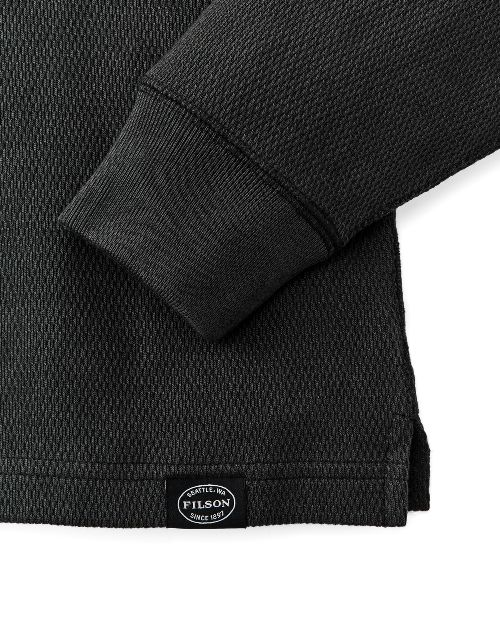 Close-up of a black textured knit garment, showing a ribbed cuff and a small oval FILSON label near the hem on a white background.