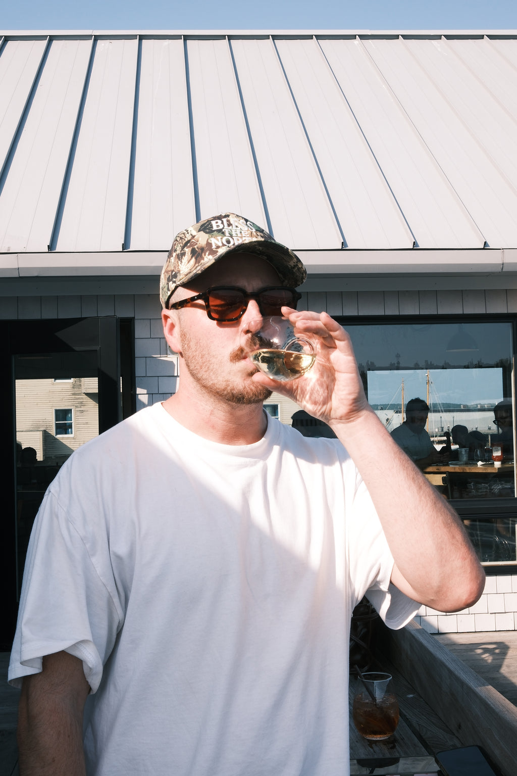 A man wearing a white t-shirt, camouflage baseball cap, and sunglasses drinks from a small glass while standing outdoors in front of a building with a metal roof.