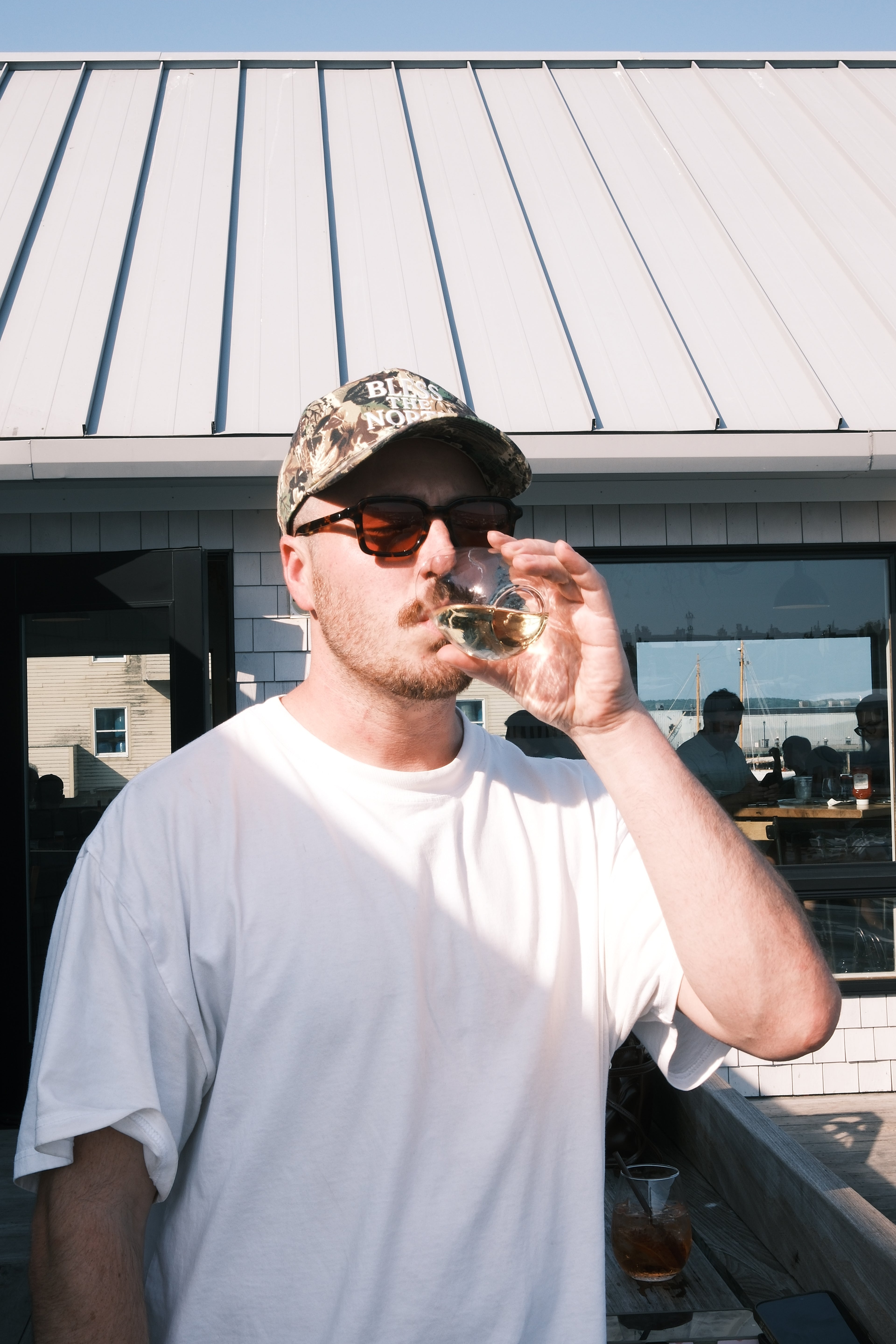 A man wearing a white t-shirt, camouflage baseball cap, and sunglasses drinks from a small glass while standing outdoors in front of a building with a metal roof.