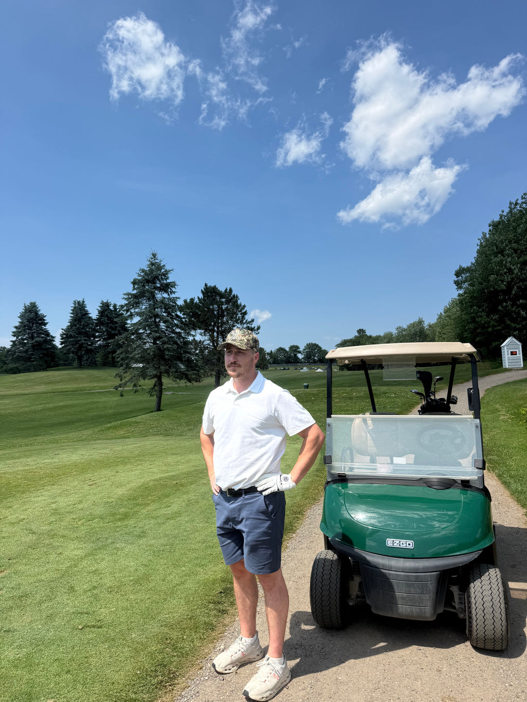 A man wearing a white polo shirt, navy shorts, and a camouflage cap stands next to a green golf cart on a golf course path under a blue sky with white clouds.