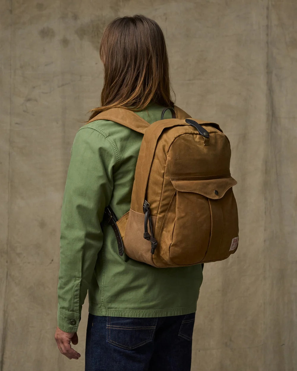 A person viewed from behind wearing a tan waxed canvas backpack over an olive green jacket and dark blue jeans, standing against a textured concrete wall.