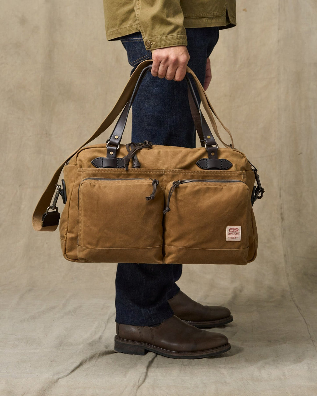 Person holding a brown leather duffel bag against a beige background