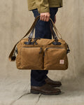 Person holding a brown leather duffel bag against a beige background