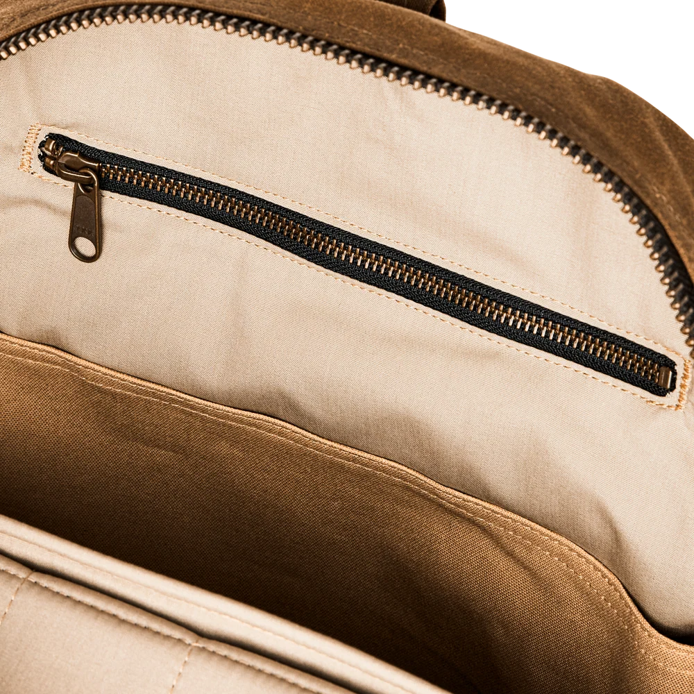 Close-up of the interior of a brown backpack showing a beige fabric pocket with a black zipper and brass zipper pull.