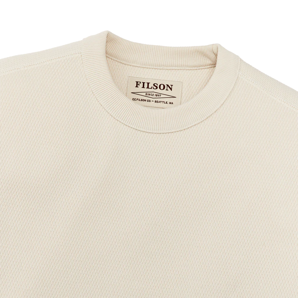 Close-up of a cream-colored crewneck sweatshirt by Filson, showing the ribbed collar and the brand label inside.