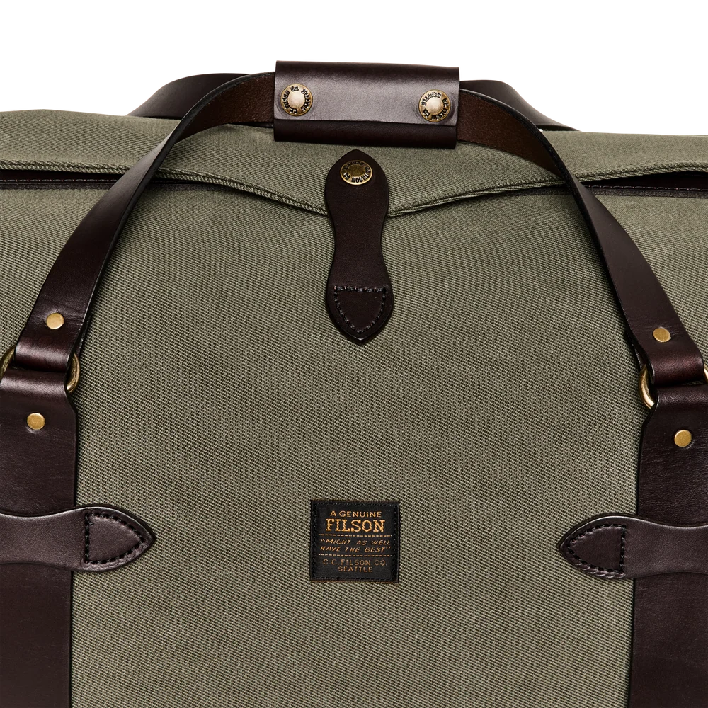 Close-up of a Filson duffel bag in olive green canvas with dark brown leather straps and hardware, featuring a visible Filson brand patch on the front.