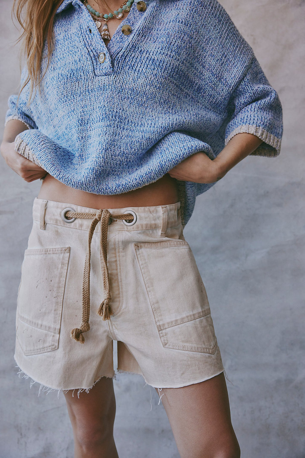 Person wearing a blue knitted poncho and beige shorts against a neutral background