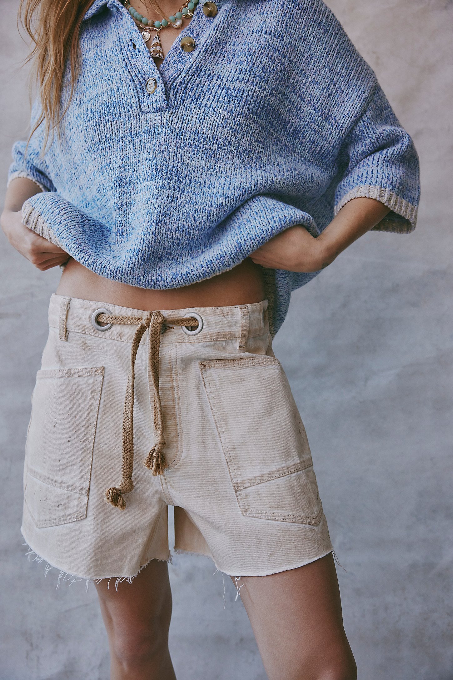 Person wearing a blue knitted poncho and beige shorts against a neutral background
