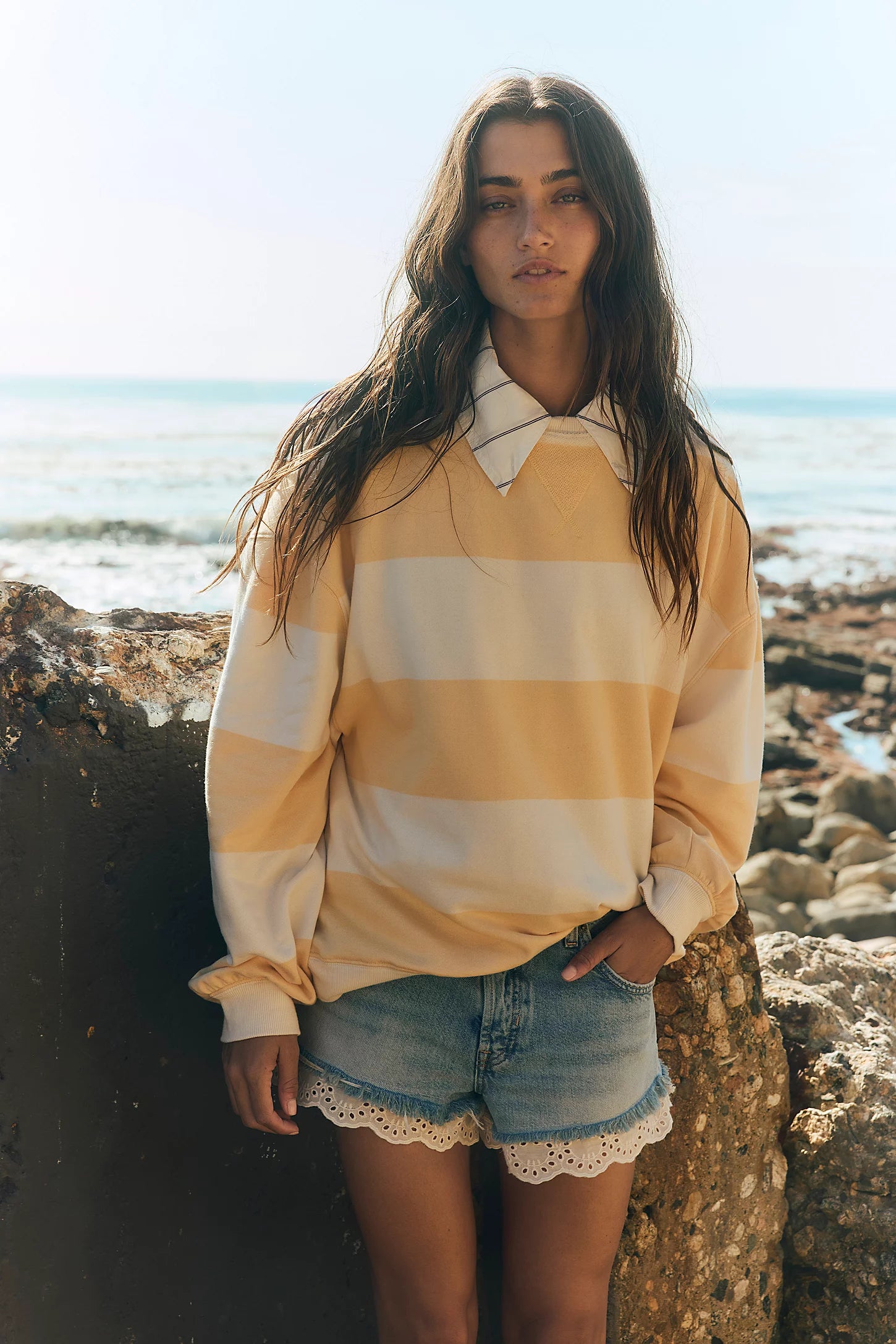 Woman wearing a yellow and white striped sweater and denim shorts standing on rocky terrain by the ocean.