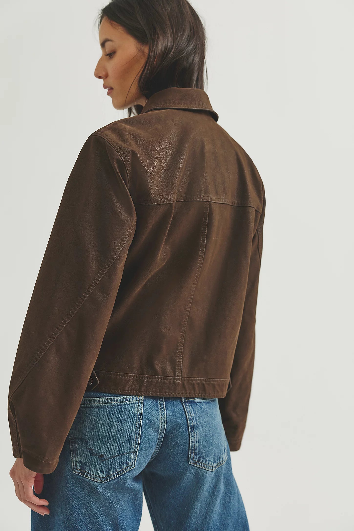 A woman shown from the back wearing a brown cropped jacket and blue jeans against a plain light gray studio background.