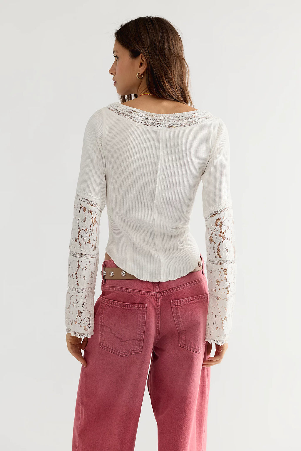 A woman shown from the back wearing a white long-sleeve top with lace detailing on the sleeves and neckline, paired with dusty rose pants, against a plain light gray studio background.