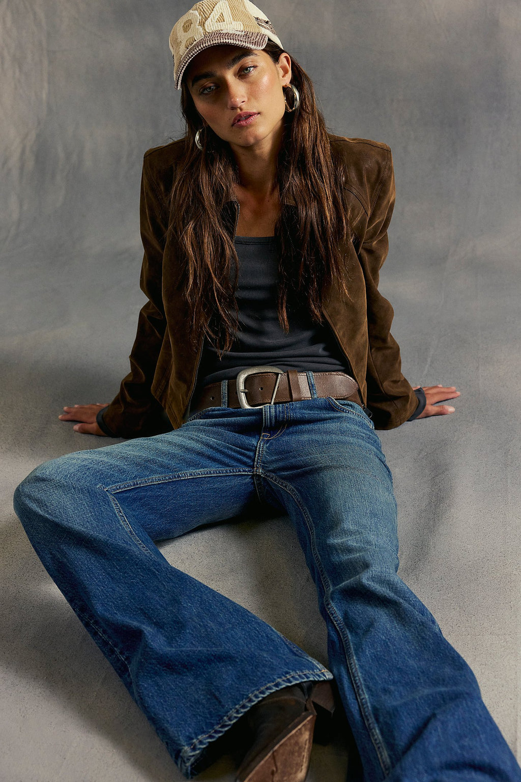 Woman wearing a brown jacket, blue jeans, and a cap sitting on a gray surface.