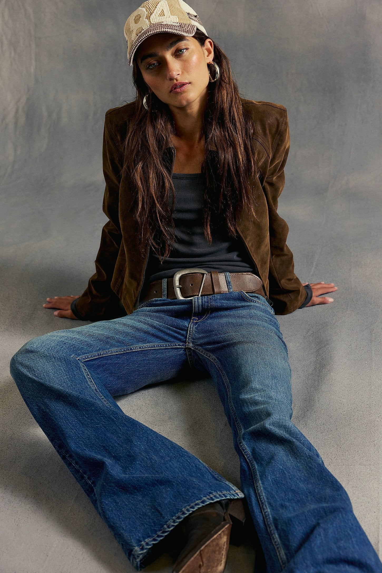 Woman wearing a brown jacket, blue jeans, and a cap sitting on a gray surface.