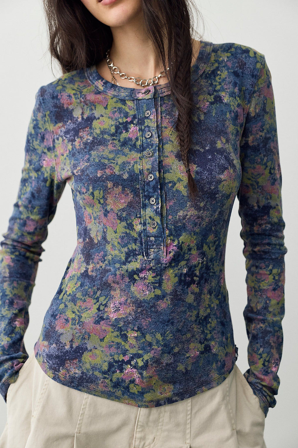 A woman wearing a long-sleeve Henley-style top with a blue, purple, green, and pink floral pattern, paired with beige pants and a chunky silver chain necklace.