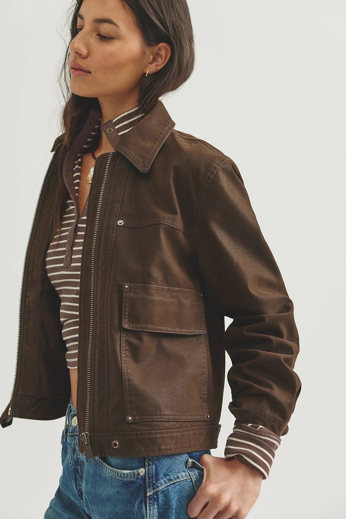 A woman wearing a brown leather jacket over a striped shirt and blue jeans, photographed against a plain light gray studio background.