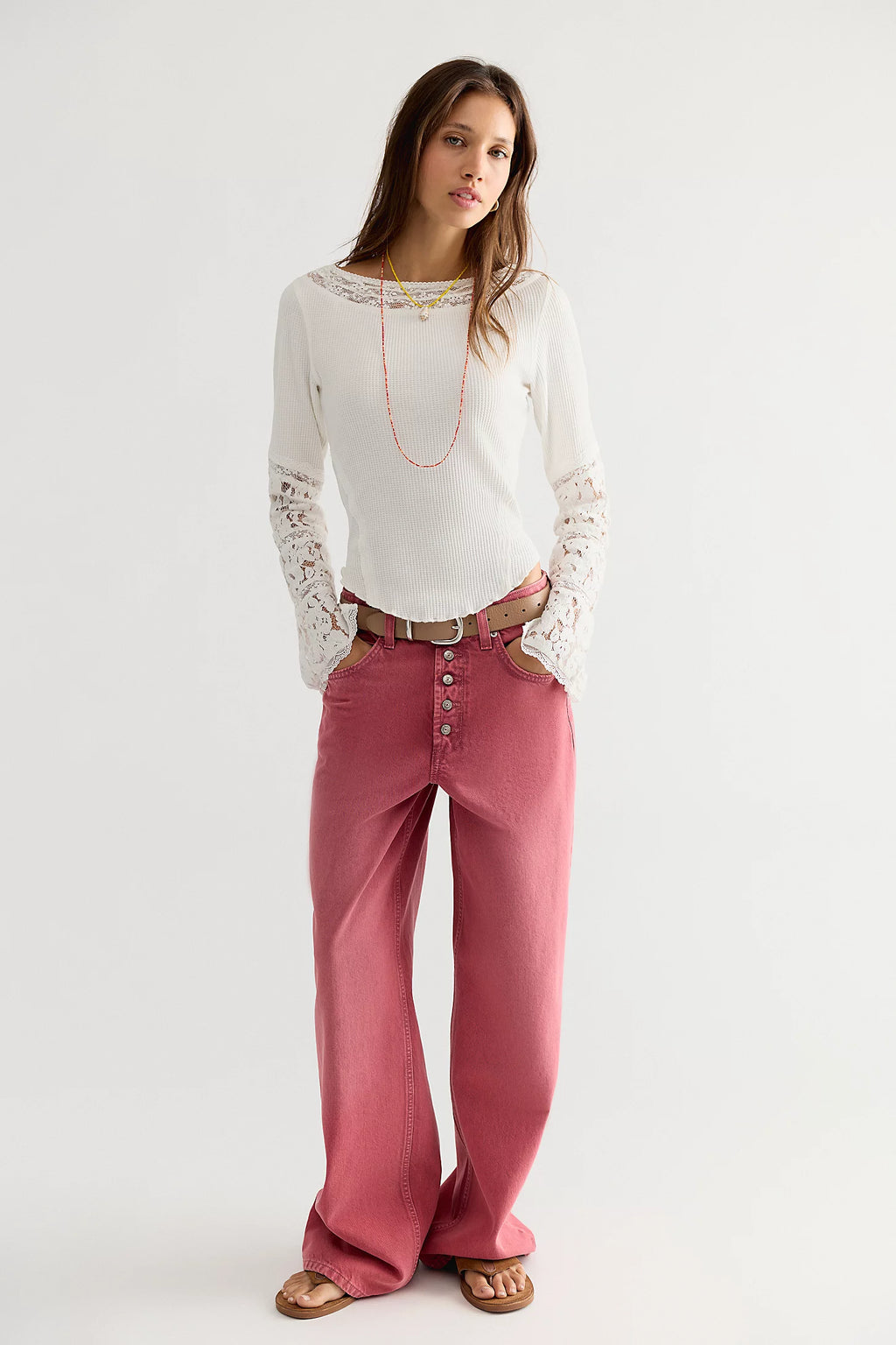 A woman modeling a white long-sleeve top with lace sleeves and pink wide-leg pants, styled with a brown belt, necklace, and sandals against a plain light gray studio background.
