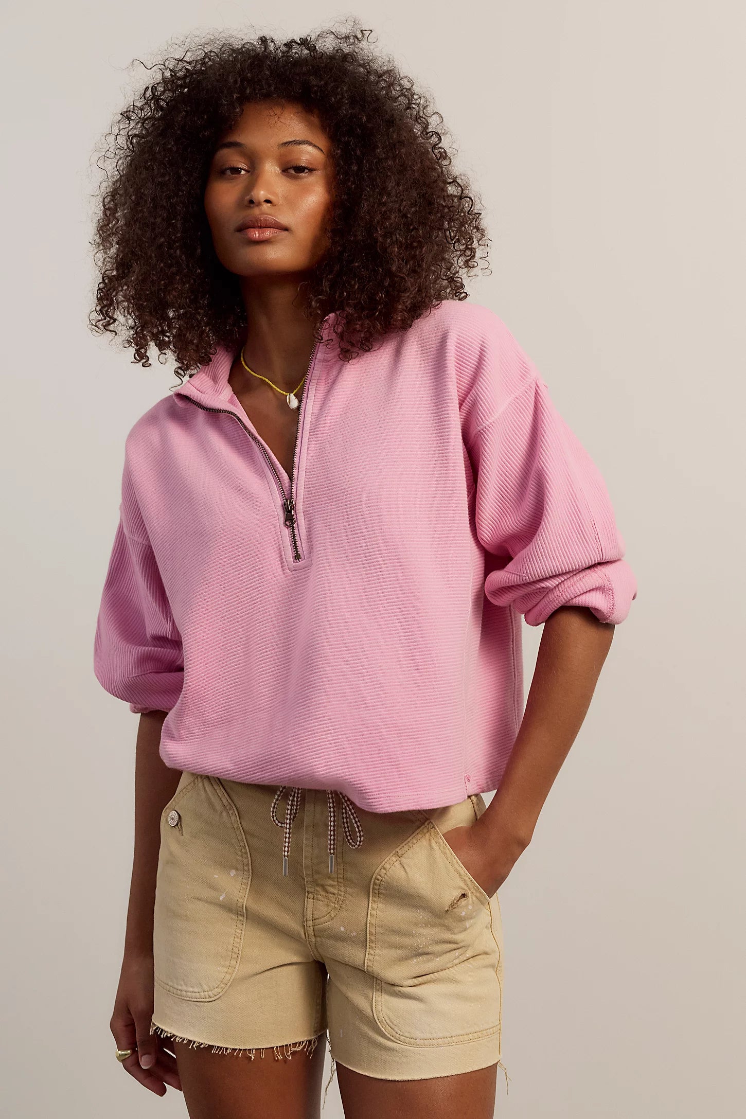 Woman wearing a pink sweater and beige shorts against a plain background