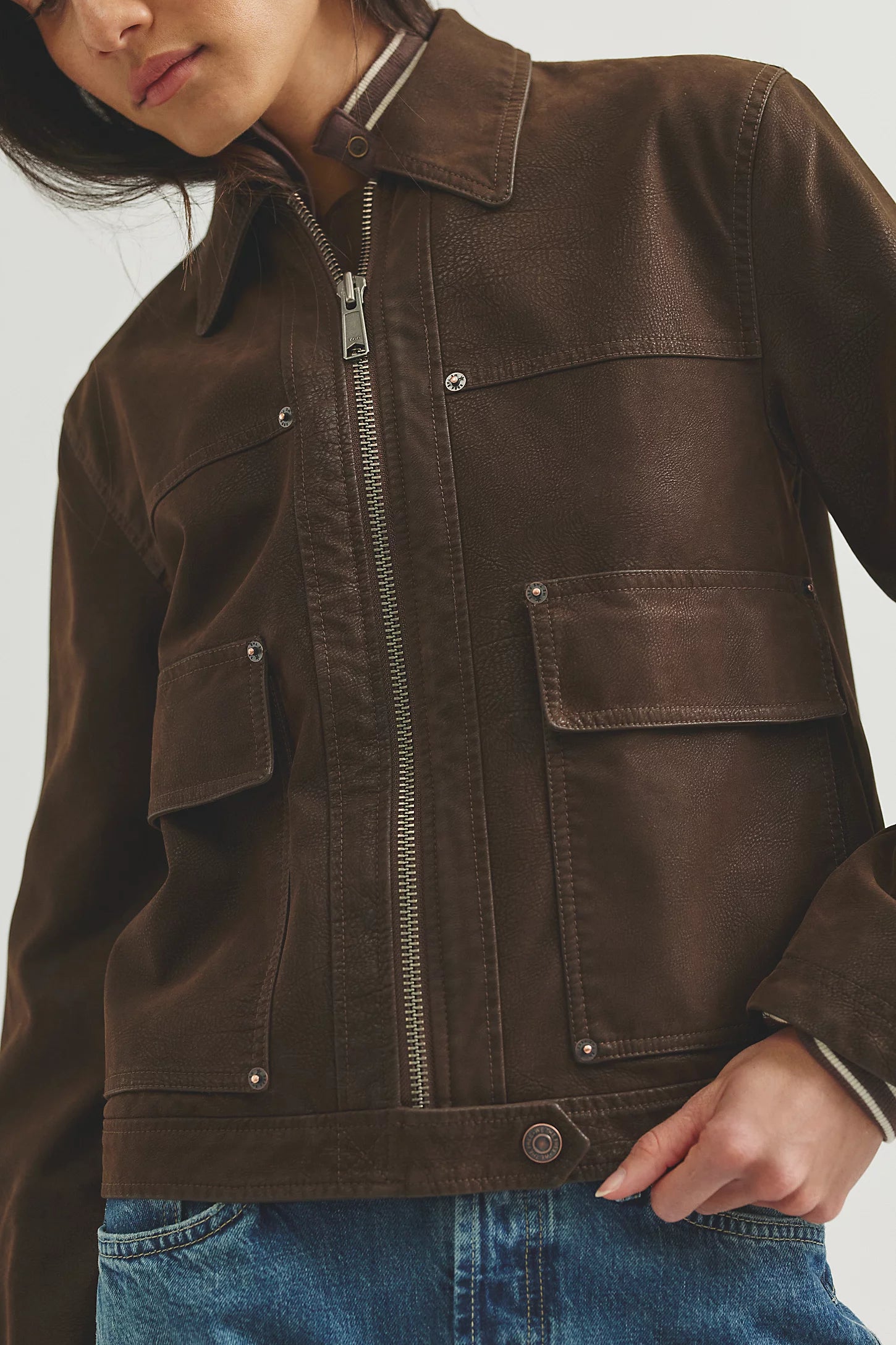A close-up of a model wearing a dark brown canvas utility jacket with a front zipper, button details, and flap pockets, paired with blue jeans.