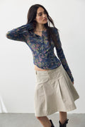 A woman modeling a sheer floral long-sleeve crop top in blue, purple, and green tones, paired with a beige pleated A-line skirt and black ankle boots, against a plain white studio background.