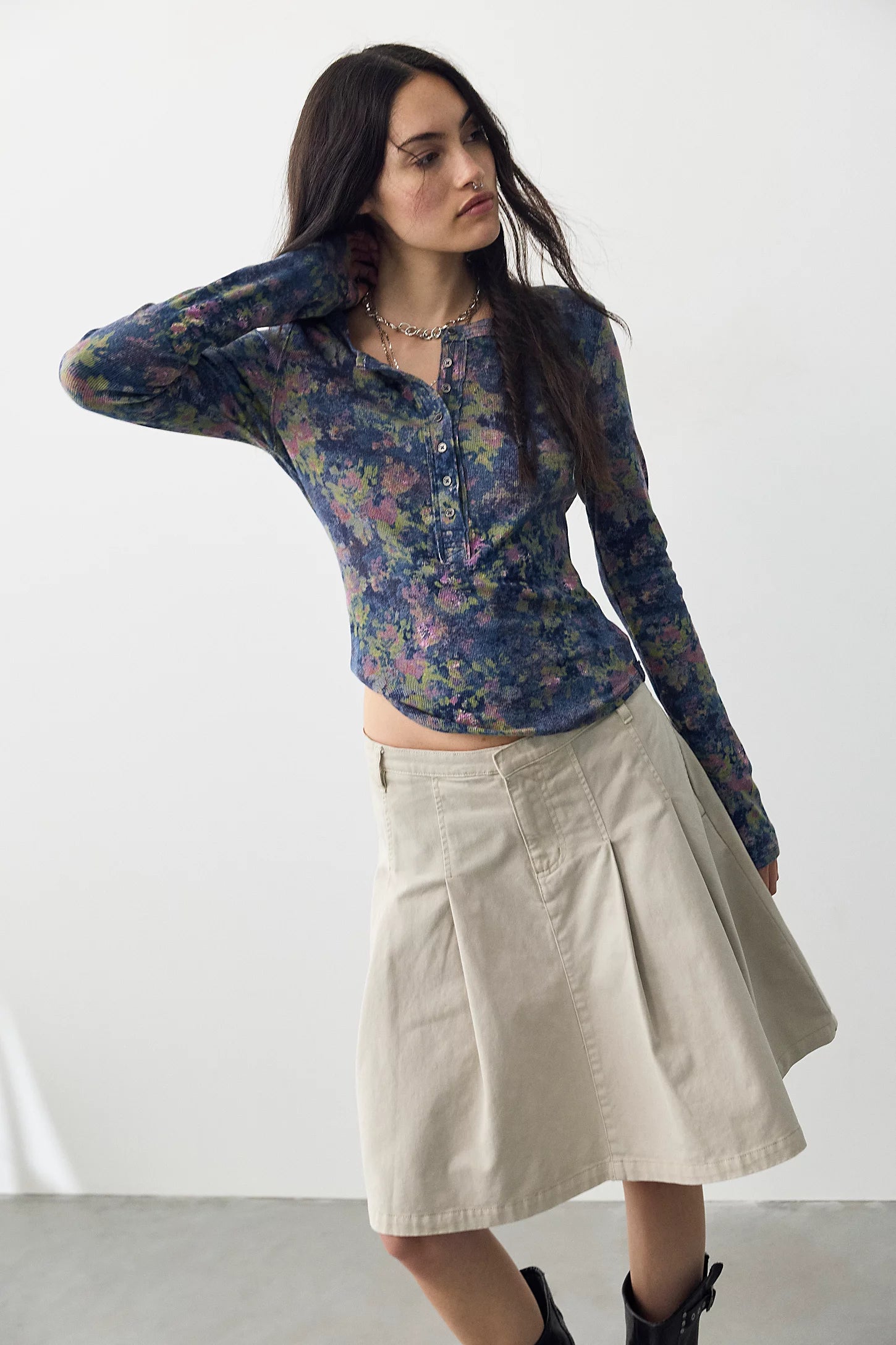 A woman modeling a sheer floral long-sleeve crop top in blue, purple, and green tones, paired with a beige pleated A-line skirt and black ankle boots, against a plain white studio background.