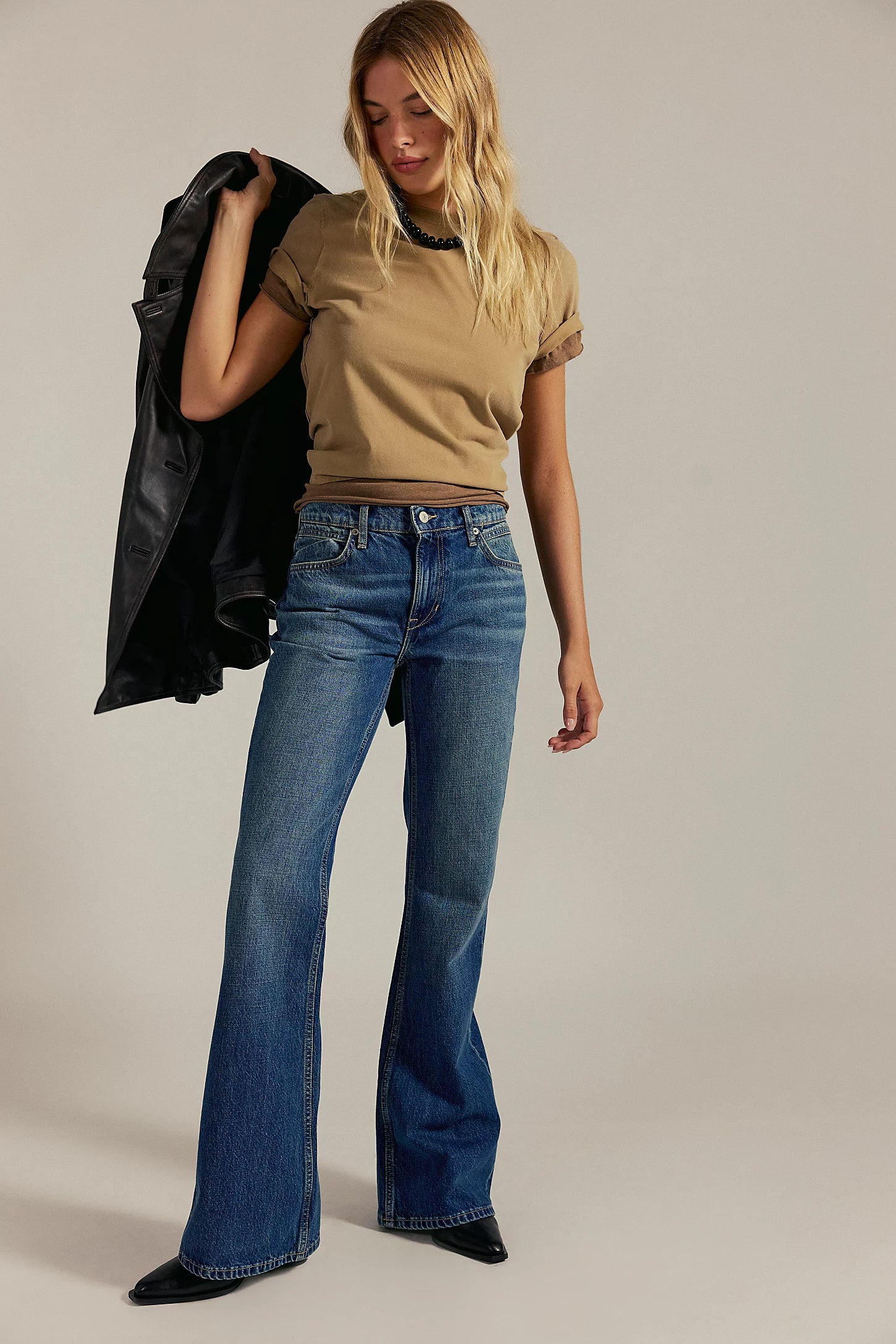 Woman wearing a beige top and blue jeans holding a black jacket against a neutral background
