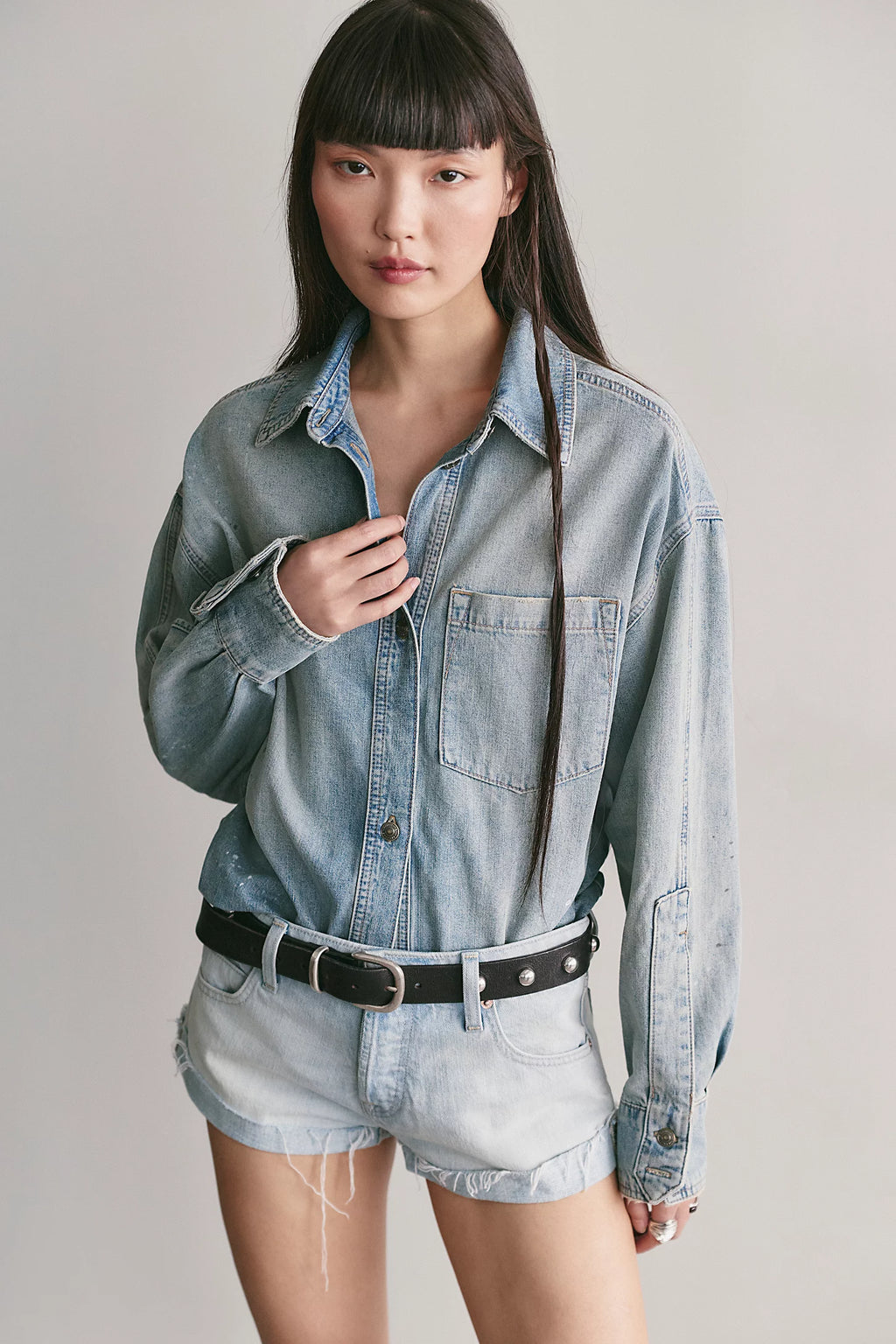 A model wearing a light-wash blue denim button-up shirt tucked into matching light-wash denim shorts, styled with a black belt, against a plain light gray studio background.