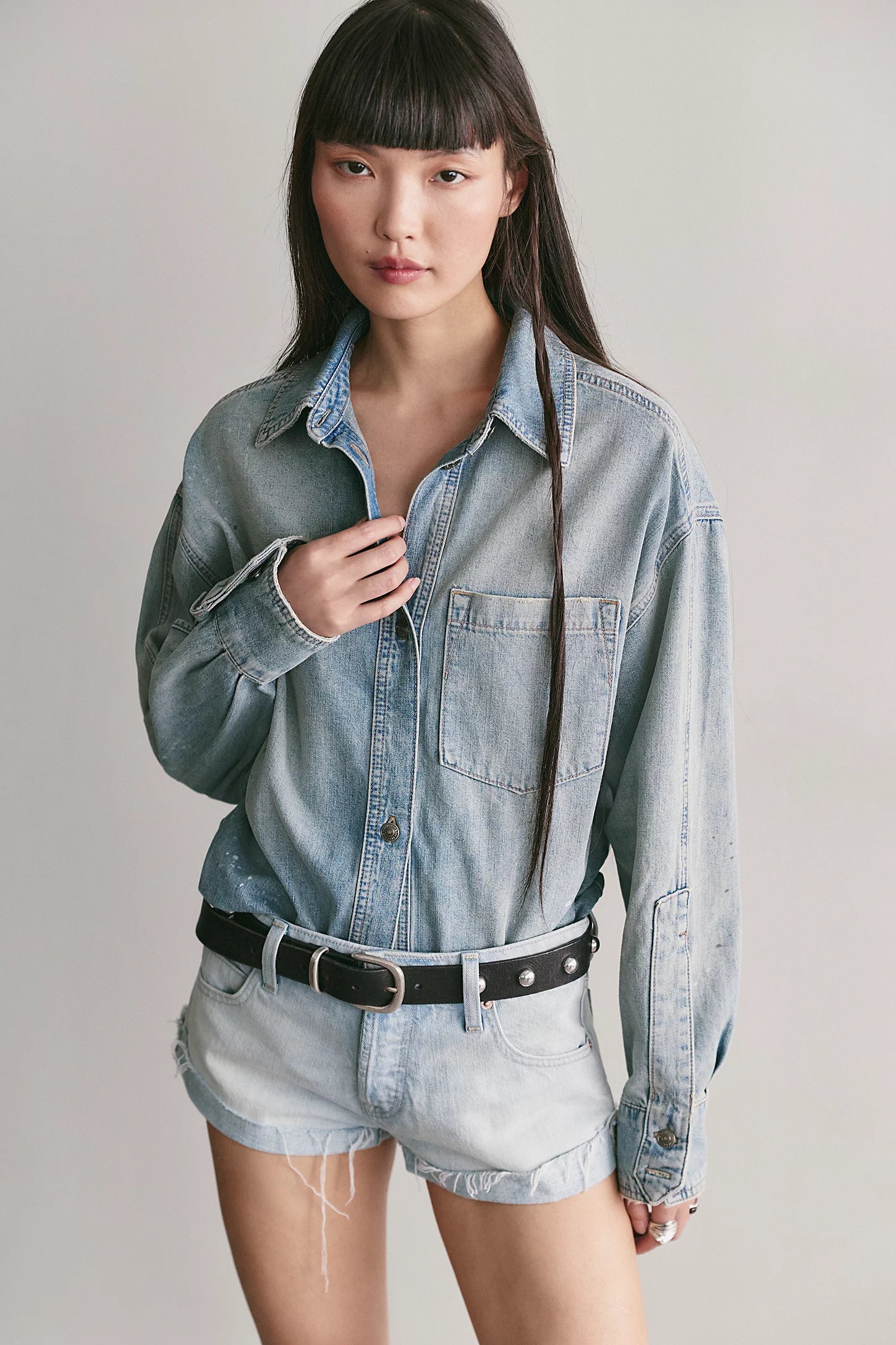 A model wearing a light-wash blue denim button-up shirt tucked into matching light-wash denim shorts, styled with a black belt, against a plain light gray studio background.