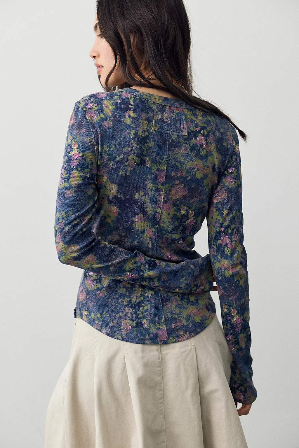 A woman modeling a sheer floral long-sleeve crop top in blue, purple, and green tones, paired with a beige pleated A-line skirt and black ankle boots, against a plain white studio background.