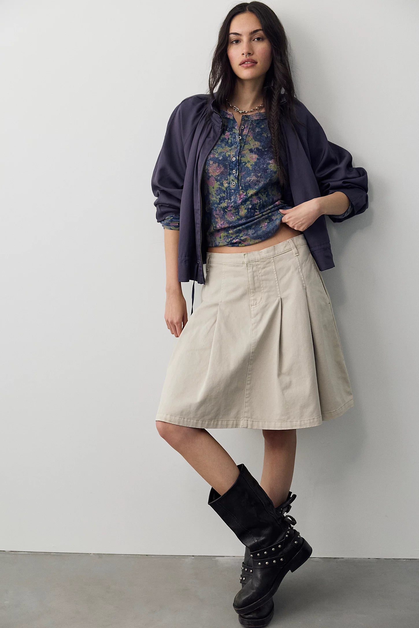 A woman models a stylish outfit consisting of a navy blue cropped jacket, a floral print crop top, a pleated beige skirt, and black studded combat boots, posing against a light gray wall.