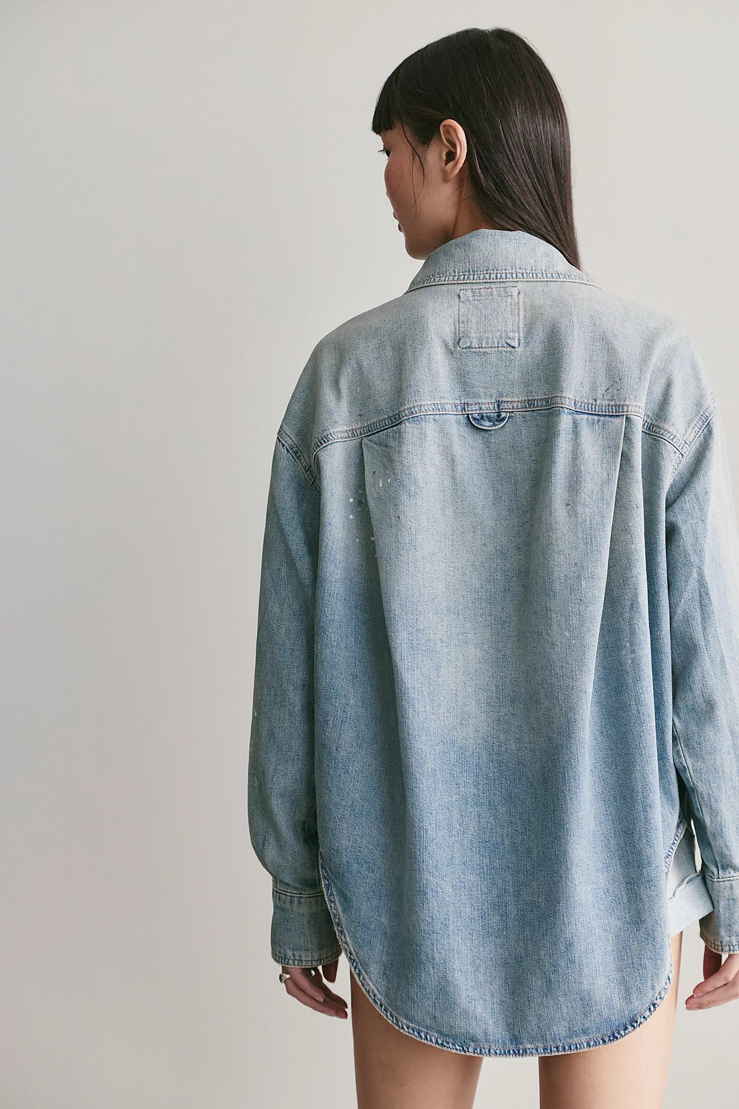 A model shown from the back wearing an oversized light-wash blue denim shirt jacket, set against a plain light gray studio background.