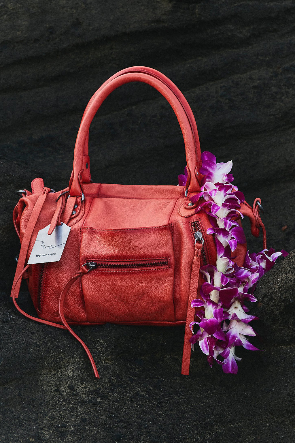 Red handbag with a tag on a dark surface, accompanied by purple flowers.