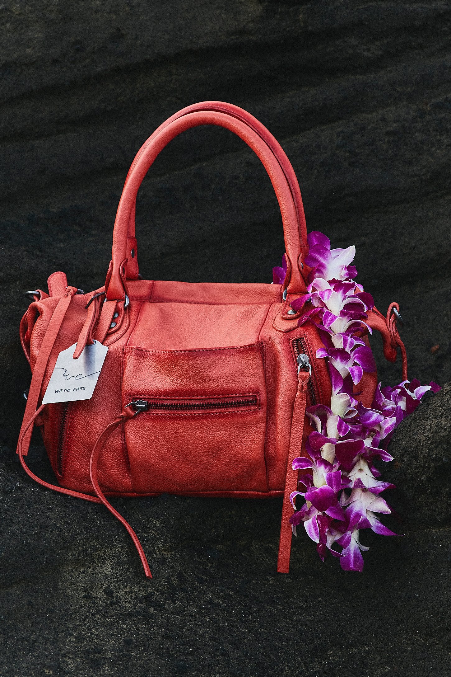 Red handbag with a tag on a dark surface, accompanied by purple flowers.