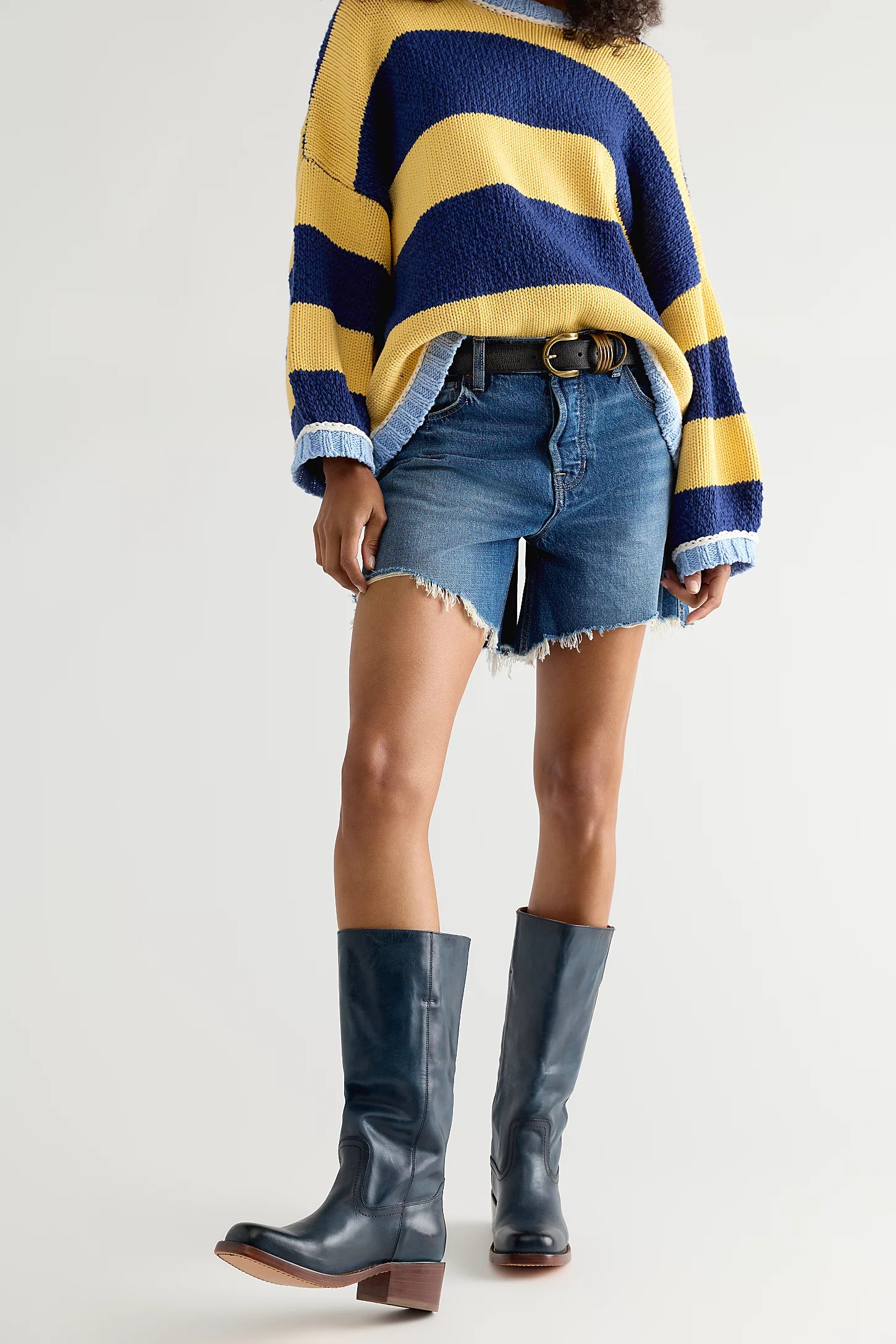 A model wearing a navy and yellow striped fuzzy knit sweater, frayed denim shorts, and dark blue leather knee-high boots, shown from the chest down against a light gray studio background.