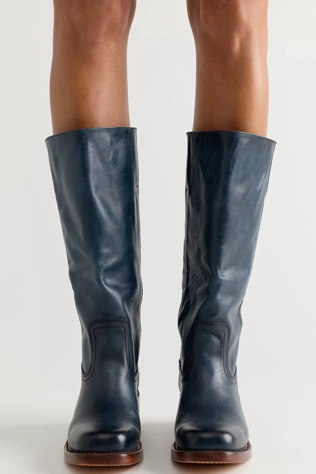 A pair of dark navy blue leather knee-high riding boots with brown soles, shown on a model's legs against a light gray studio background.