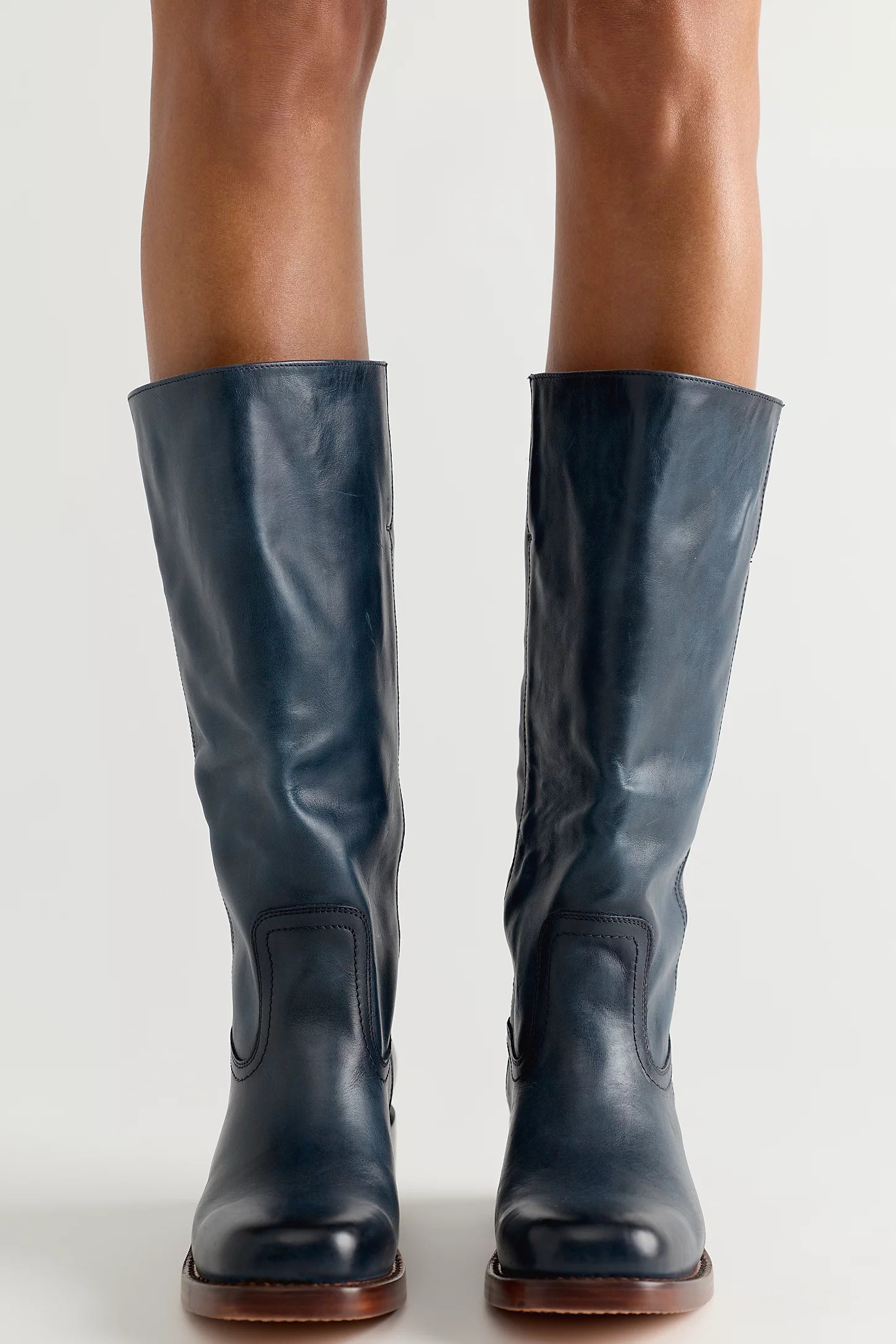A pair of dark navy blue leather knee-high riding boots with brown soles, shown on a model's legs against a light gray studio background.