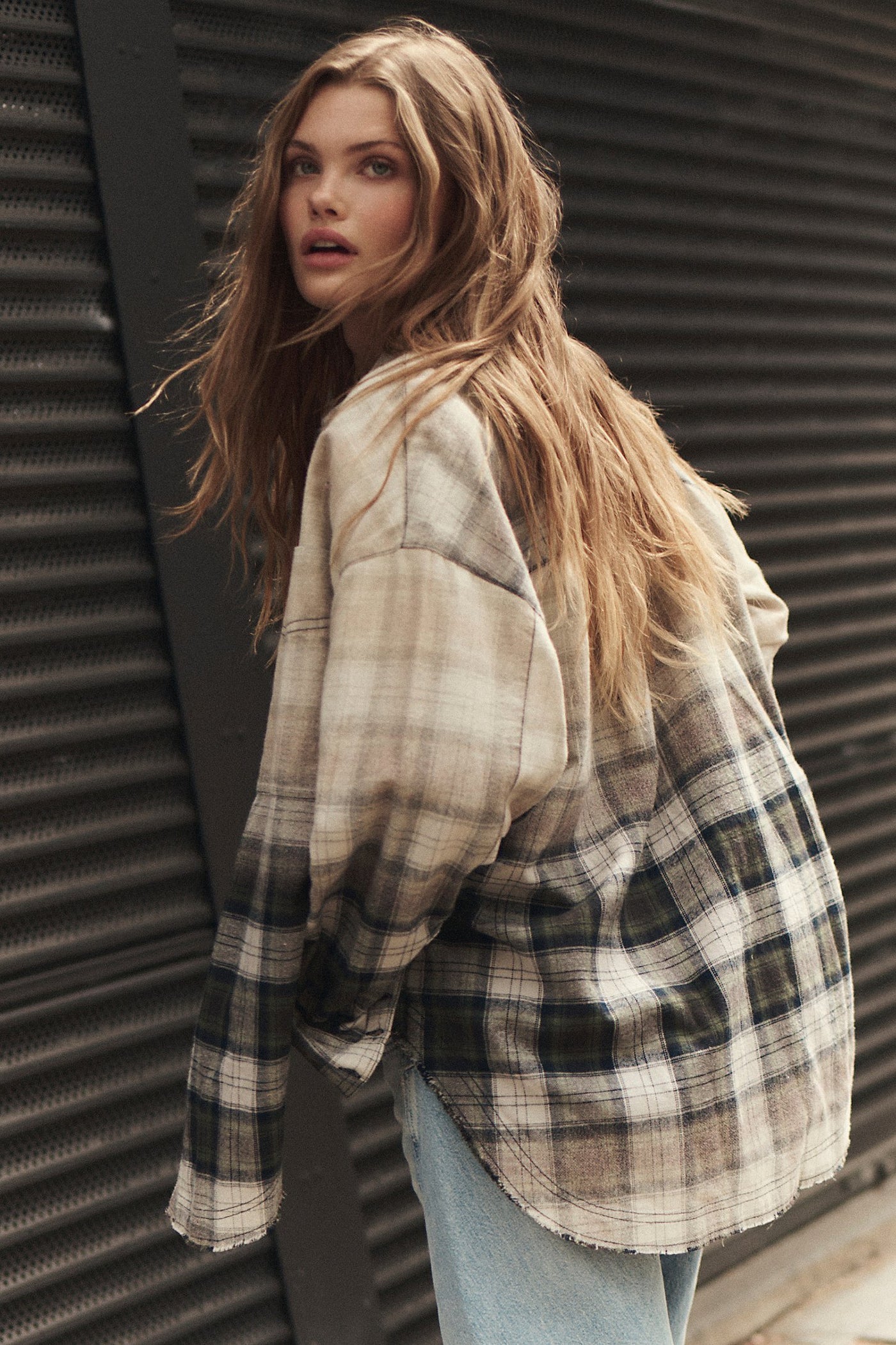 A woman wearing an oversized plaid flannel shirt with a beige, brown, and black pattern, paired with light blue jeans, standing in front of a dark corrugated metal wall.