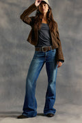 Woman wearing a brown jacket, gray shirt, blue jeans, and a cap against a gray background