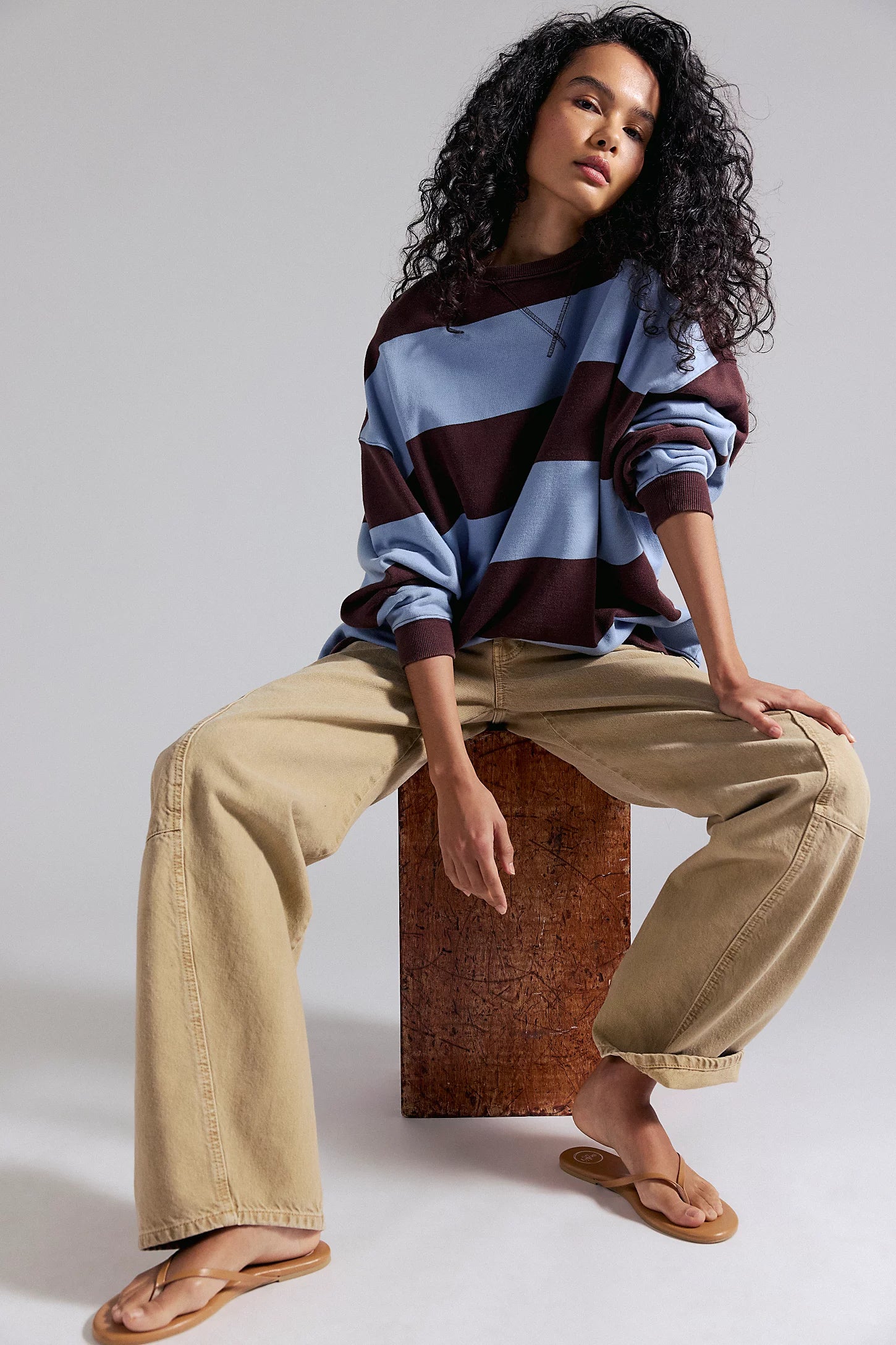 Person wearing a striped sweater and beige pants sitting on a wooden block against a plain background