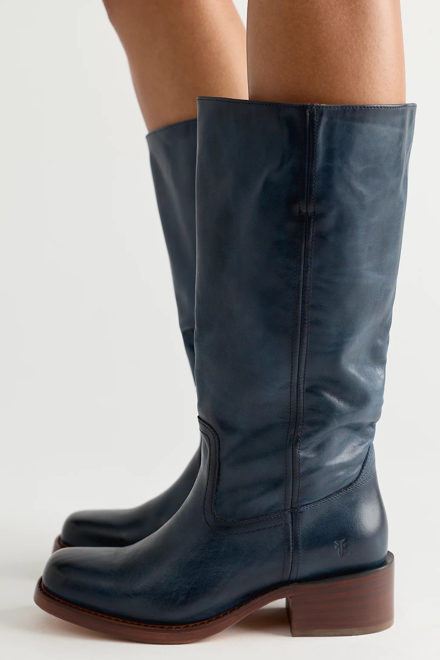 A pair of dark navy blue leather knee-high boots with brown stacked block heels, shown on a model's feet against a light gray studio background.