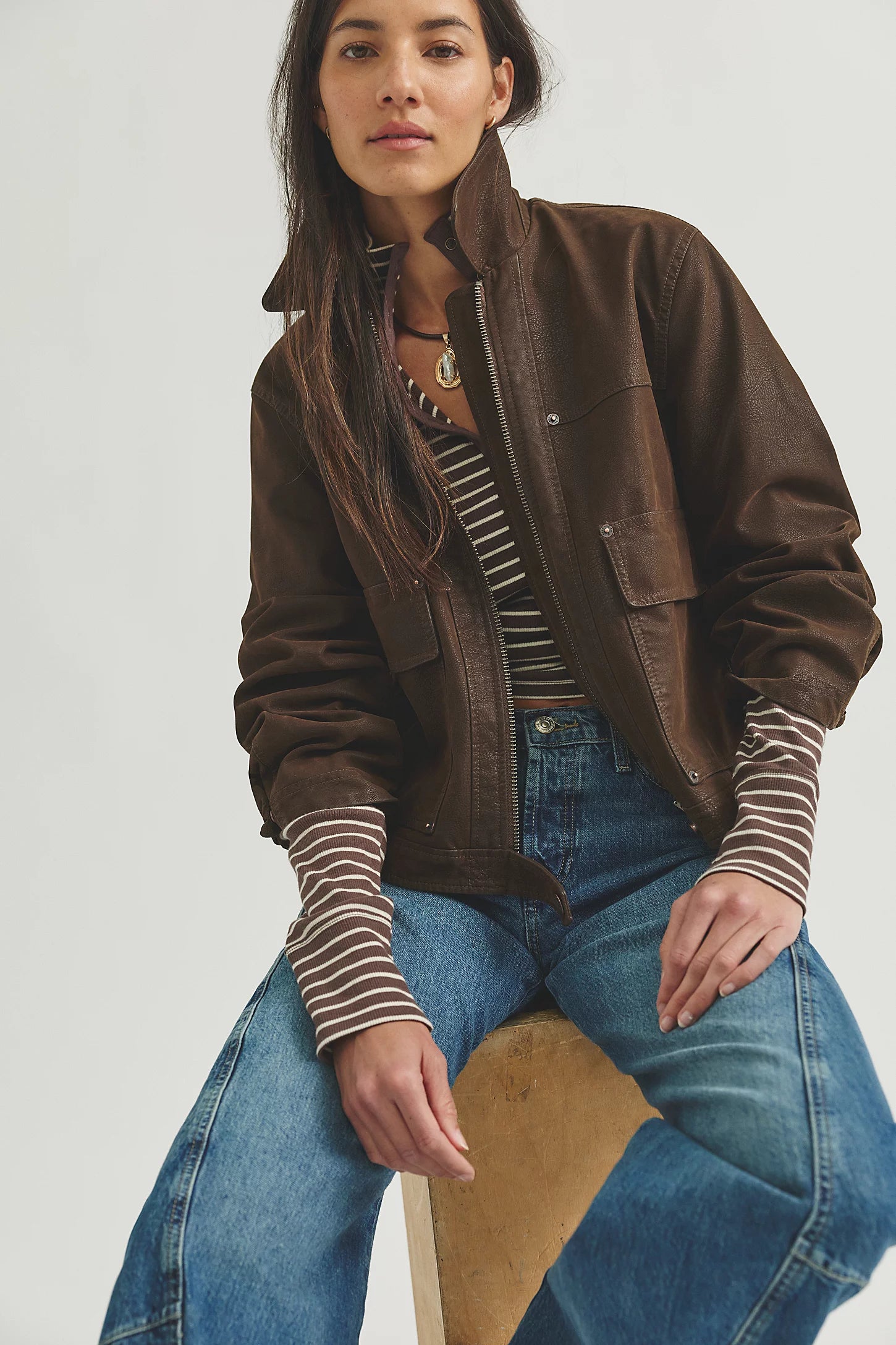 A woman wearing a brown suede-like jacket over a striped long-sleeve top and blue jeans, seated on a wooden stool against a light gray studio background.