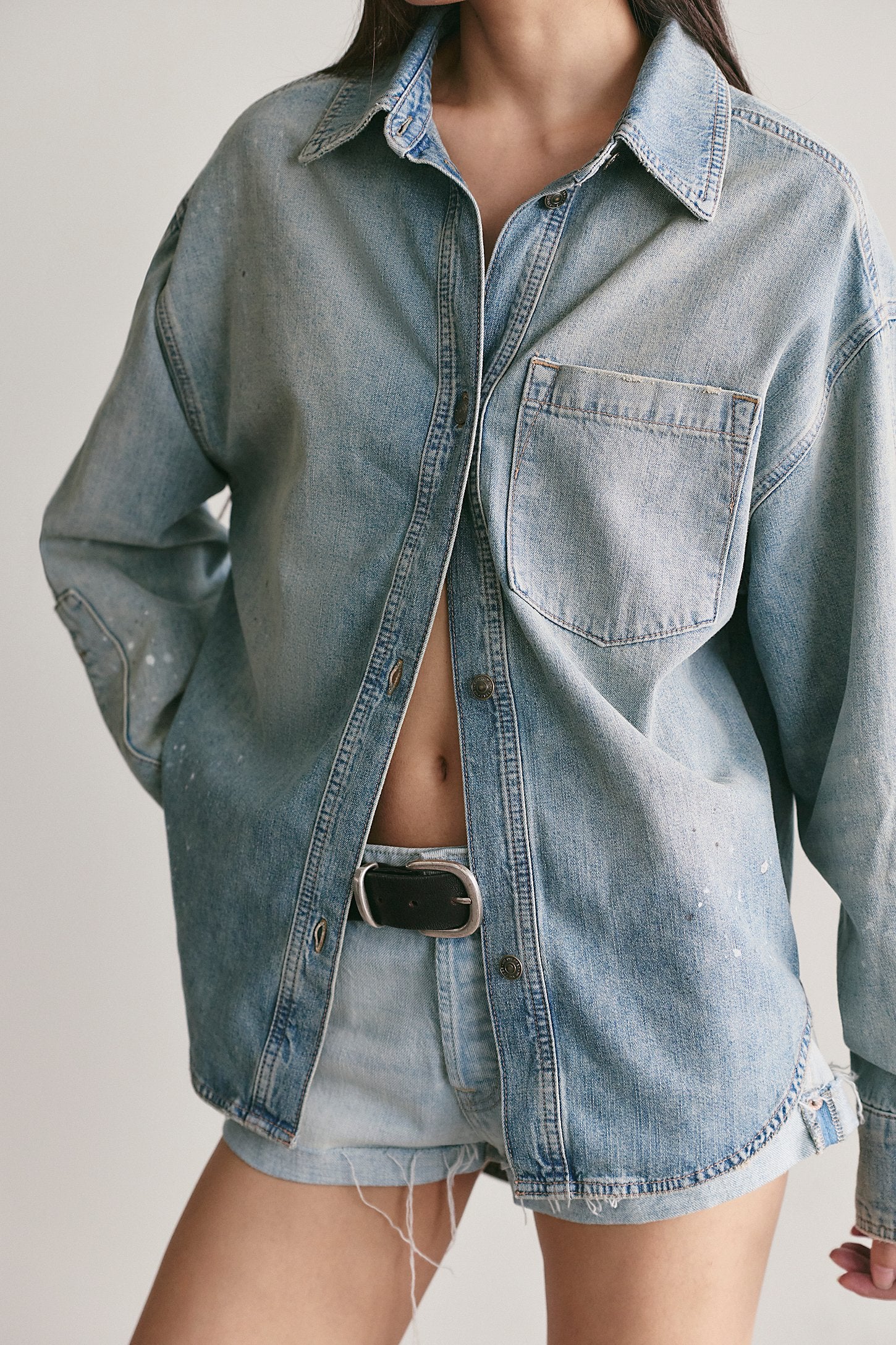 A person wearing an oversized light-wash blue denim button-up shirt with a chest pocket, paired with matching denim shorts and a black belt, against a plain light background.