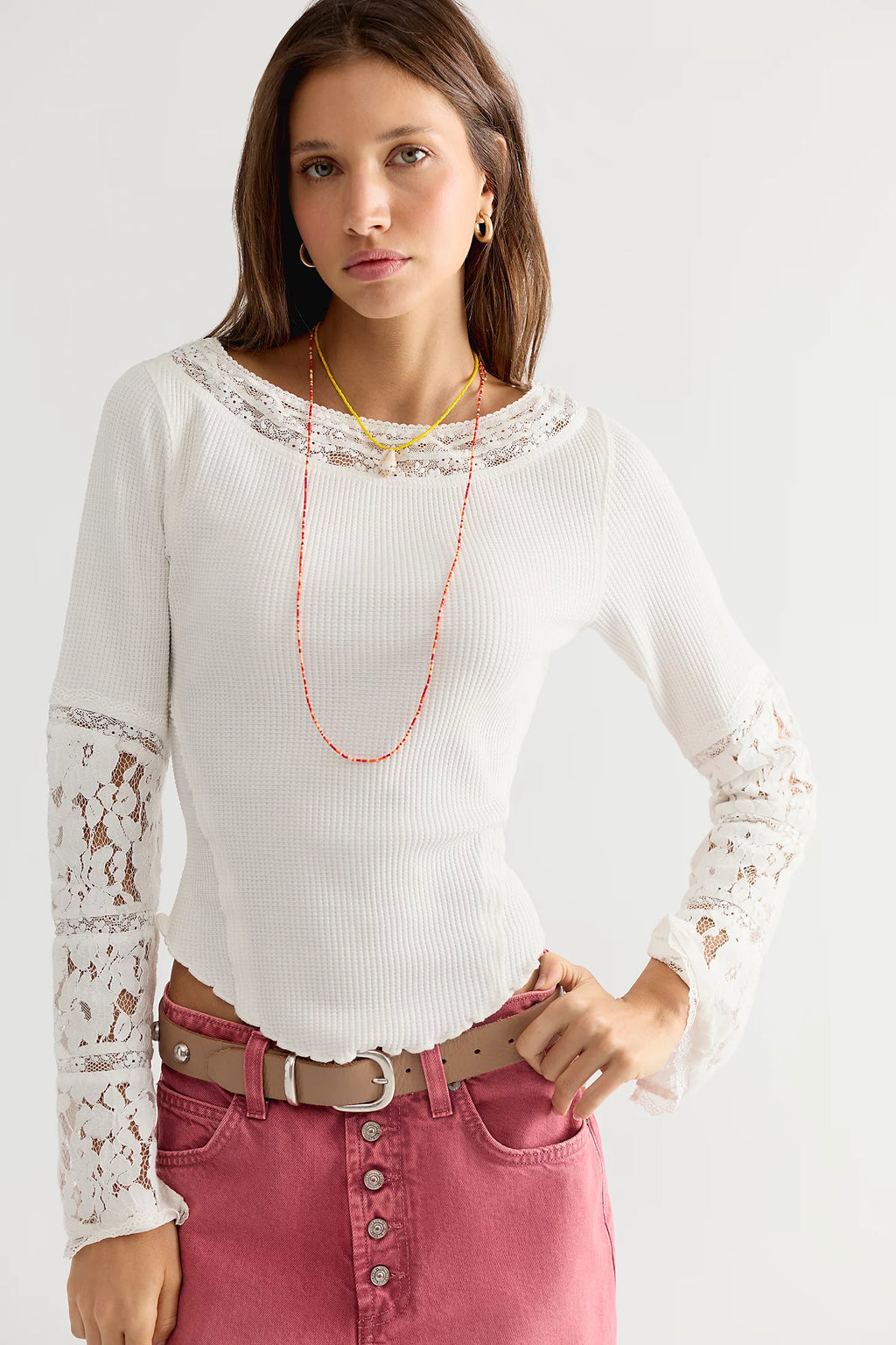 A woman wearing a white long-sleeve ribbed knit top with lace trim at the neckline and lace inserts on the sleeves, paired with pink jeans and layered necklaces, against a plain light background.