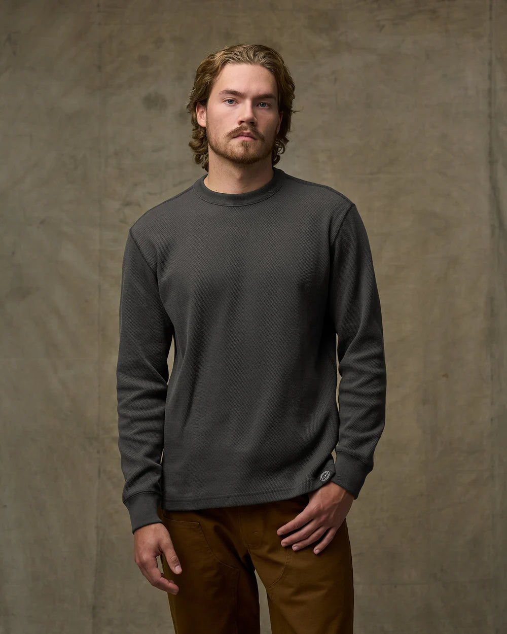 A man wearing a dark charcoal gray long-sleeve crewneck shirt and brown pants, standing against a textured brown studio backdrop.