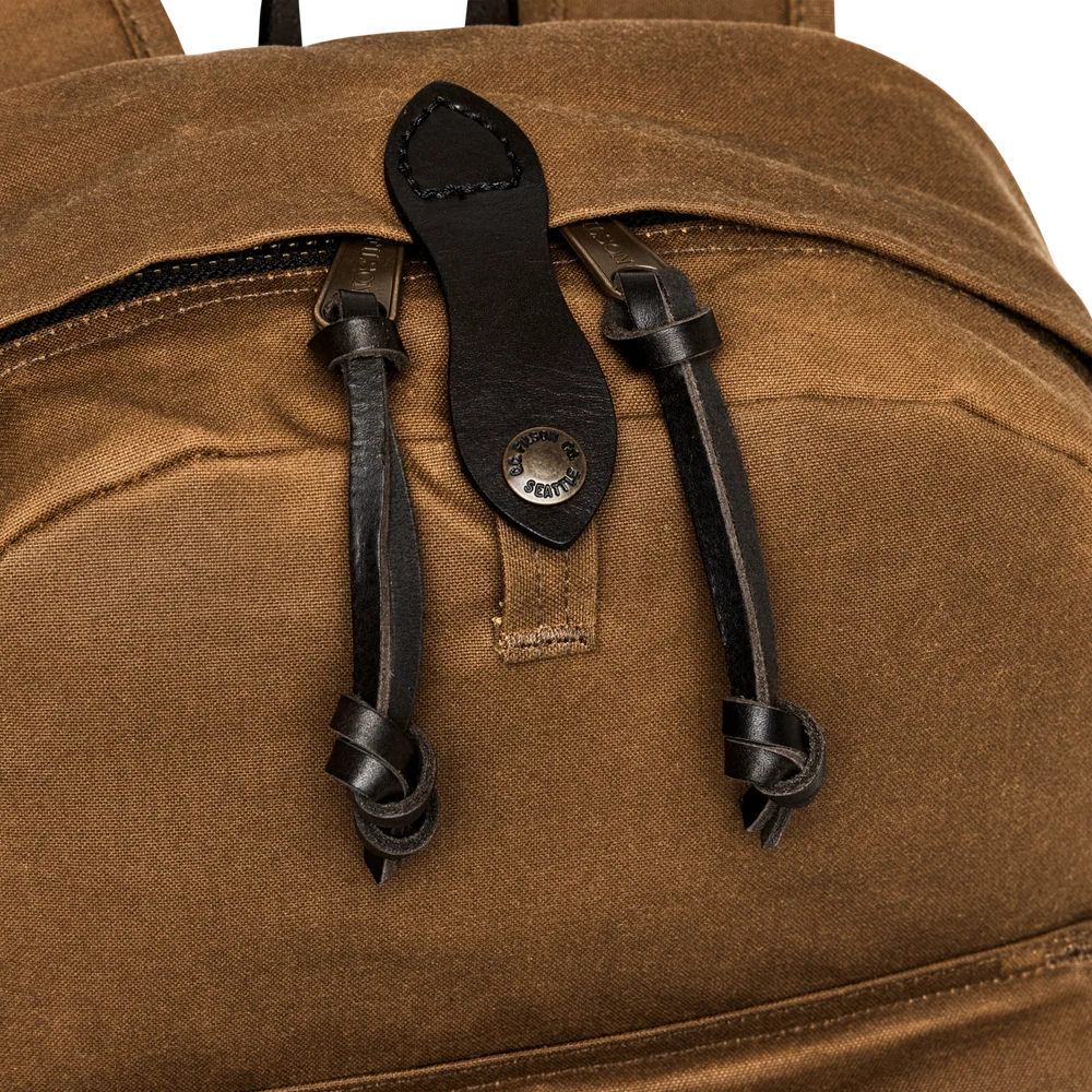 Close-up of a brown canvas backpack with black leather zipper pulls and a metal brand badge on a white background.
